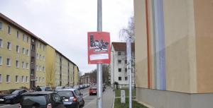 Plakatieren - Werbung am Lichtmast auf einer Straße, im Hintergrund Häuserzüge