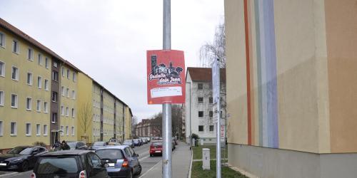 Plakatieren - Werbung am Lichtmast auf einer Straße, im Hintergrund Häuserzüge