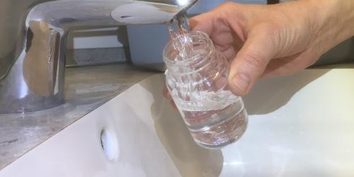 Wasser läuft aus dem Wasserhahn in ein steriles Probennahmeglas zur Überprüfung der mikrobiologischen Trinkwasserqualität.