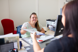 Eine Frau sitzt an einem Schreibtisch an einem Rechner und übergibt einer anderen Frau, die man von hinten seitlich sieht, ein Dokument.