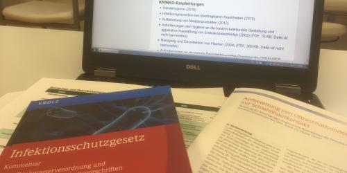 Arztzimmer mit Tisch auf diesem sich ein Laptop und ein Gesetzesbuch - Infektionsschutzgesetz befinden